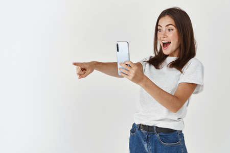 Excited young woman taking picture on mobile phone, pointing left at product and smiling, recording video on smartphone, white backgroundの写真素材
