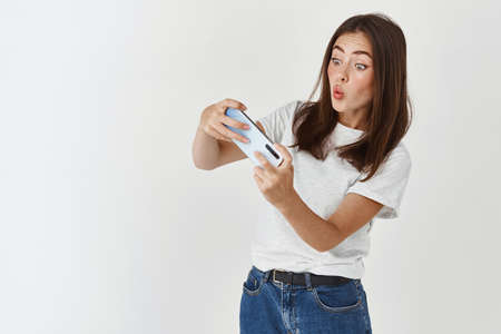 Excited brunette woman tilting body while playing racing video game on smartphone, looking amused,...
