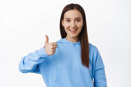 Very good. Image of young woman in hoodie showing thumb up in approval, like and agree, praise good...