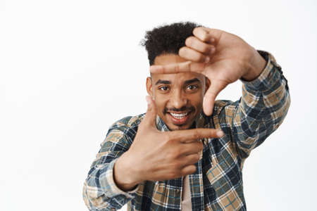 Creative african american male student, guy looking through finger frames and smiling amused, capture moment, taking photo camera, imaging something, white backgroundの写真素材