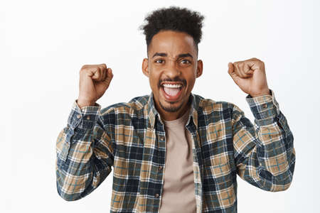 Young cheerful african american man scream in support, shouting like sports fan, shaking clenched...