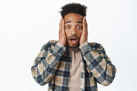 Portrait of shocked african american man gasping speechless, holding hands on face and staring...