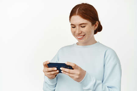 Young teen redhead girl playing video games on her phone, watching video live stream on cellphone,...