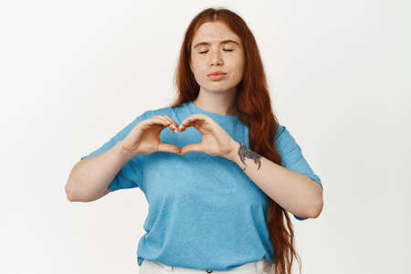 Cute redhead girl, pucker lips for kiss and show heart I love you sign, adore or like someone, show her affection or sympathy, dreaming of romantic moment, standing against white backgroundの写真素材