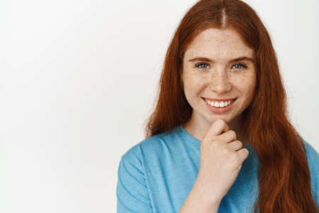 Close up portrait of smiling redhead girl, looking thoughtful, touch chin and gazing at camera, ponder smth, listening with interest, standing in tshirt against white backgroundの写真素材