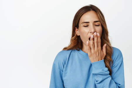 Image of tired adult woman, mother yawning and covering opened mouth with hand, exhausted middle aged female, waking up early, standing with fatigue against white backgroundの写真素材