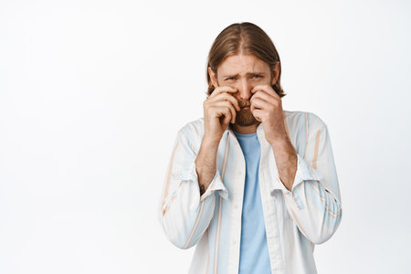 Portrait of silly and sad blond guy feeling timid, adult man crying and looking upset, wiping tears and rub eyes depressed, standing against white backgroundの写真素材