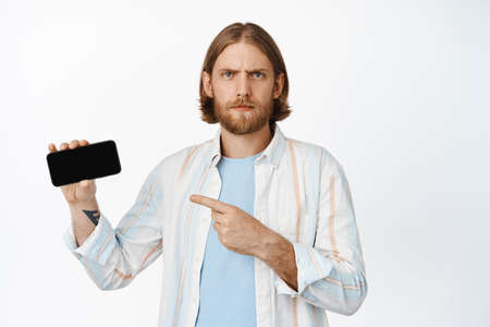 Image of confused, blond guy frowning, pointing at mobile phone, horizontal screen with concerned face, proble with app, standing against white backgroundの写真素材