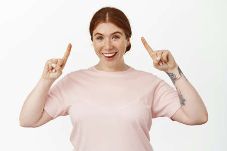 Excited redhead woman pointing fingers up at sale promo, showing best deal, offer in store, smiling...