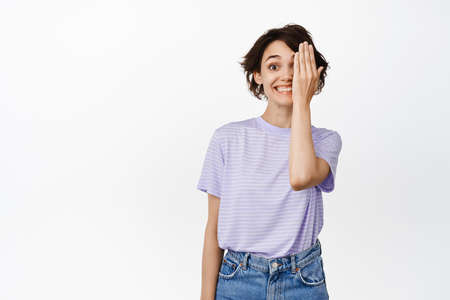 Happy beautiful girl hide one side of face, looking at camera excited, smiling, cover eye, standing...