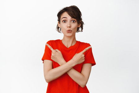 Cute young woman pointing sideways, showing left and rights sides copy space, two choices, standing in red t-shirt over white backgroundの写真素材