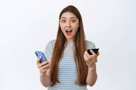 Excited and amazed young woman holding credit card and mobile phone, surprised reaction to special...