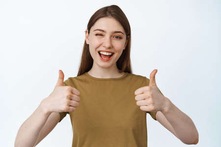 Beautiful girl winking and smiling, showing thumbs up to compliment product, praise quality,...