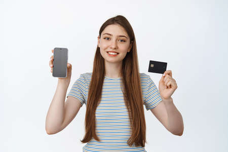 Smiling young woman showing phone screen and credit card, mobile banking and shopping concept, white...