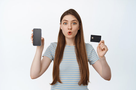 Excited and surprised girl shows credit discount card and mobile phone screen, smartphone app...