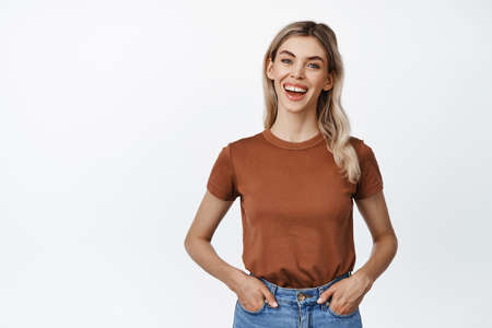 Smiling blond girl looks enthusiastic at camera, wears casual t-shirt with jeans, stands in relaxed pose against white backgroundの写真素材
