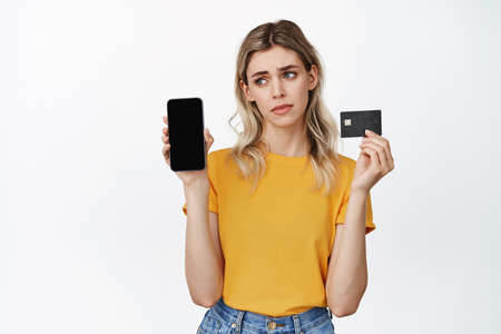 Disappointed girl shows empty smartphone and credit card, sulking sad, standing against white backgroundの写真素材