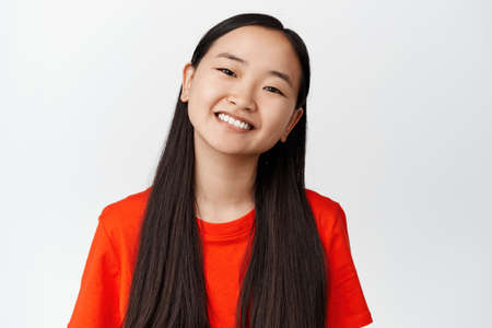 Happy asian woman smiling white teeth, looking happy and laughing, standing in red t-shirt over white backgroundの写真素材