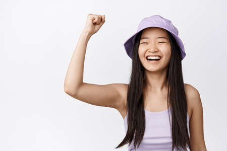 Cheerful asian girl in bucket hat celebrating, raising fist up for support, being proactive, chanting and smiling, standing over white backgroundの写真素材