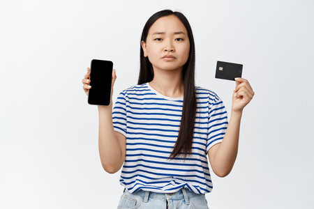 Sad frowning asian girl showing mobile phone empty screen and credit card, problem with payment...