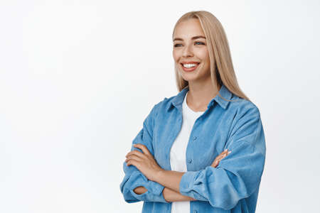 Portrait of attractive blond woman looking hopeful aside at logo, cross arms over chest and smiling,...