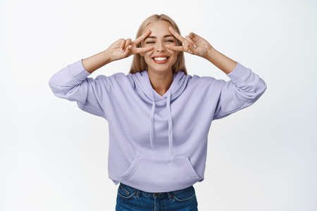 Beautiful blond girl showing positivity and joy, smiling and making peace v-signs on eyes, standing...