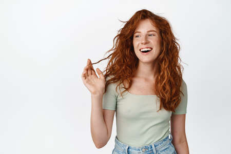 Stylish modern girl with curly red hair and freckles, playing with curls, laughing and smiling, standing in relaxed pose in t-shirt, white backgroundの写真素材