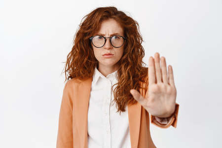 Angry redhead trainee, saleswoman showing stop, no gesture and frowning, rejecting something, declining, standing in suit and glasses against white backgroundの写真素材