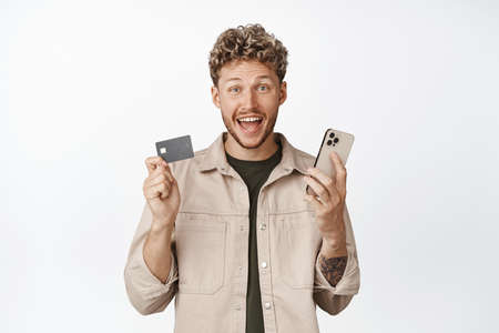 Excited young man winning money on app, holding credit card and mobile phone, shopping online,...