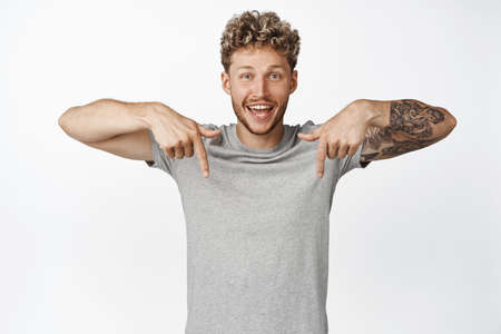 Portrait of handsome smiling blond man pointing fingers down, showing advertisement, making announcement below, standing over white backgroundの写真素材