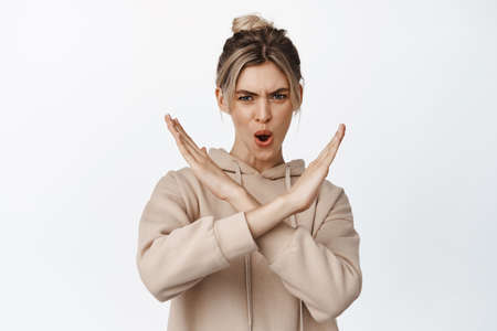 Blond young woman shows cross, stop gesture and saying no with angry and determined face expression,...