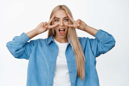 Excited young funny girl shows peace, v-sign gestures over eyes, screaming amazed and looking at...