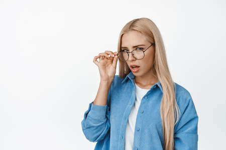 Portrait of suspicious blond woman squinting, taking off glasses and staring doubtful at camera, confused by something strange, standing over white backgroundの写真素材
