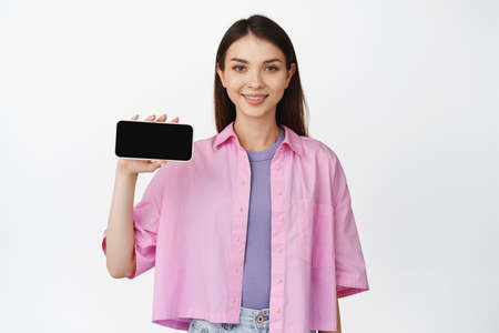Smiling beautiful girl showing smartphone screen, holding mobile phone horizontally, standing over...