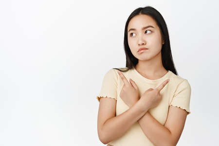 Indecisive brunette girl makes tough decision, pointing sideways and looking confused, picking from choices, standing over white backgroundの写真素材