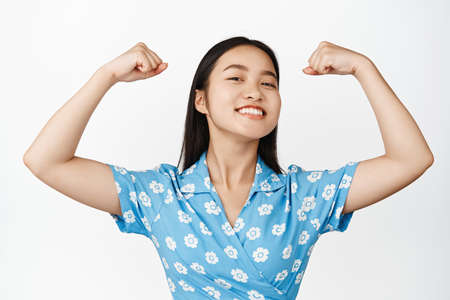 Close up of asian smiling woman feeling strength and power, flexing biceps and looking proud, showing her strong muscles, standing over white backgroundの写真素材