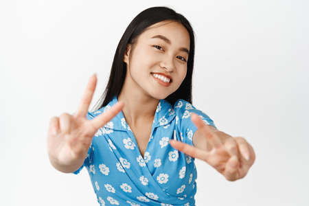 Beautiful asian female model showing peace v-sign gestures and smiling at camera, wearing blue dress, standing over white backgroundの写真素材