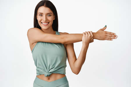 Smiling sportswoman stretching arm, showing fitness exercises. Female athlete doing warm up before...