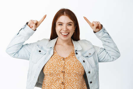 Cheerful young redhead woman pointing at black friday sale, showing advertisement up, standing over white backgroundの写真素材