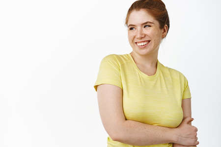 Close up of carefree redhead woman in fitness t-shirt, looking away and smiling, workout in gym, standing over white backgroundの写真素材