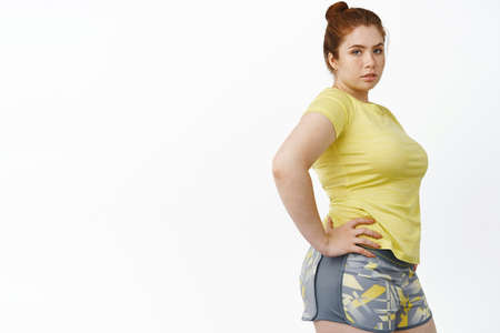 Portrait of redhead plus size woman in sport clothing, workout in gym, doing fitness exercises,...