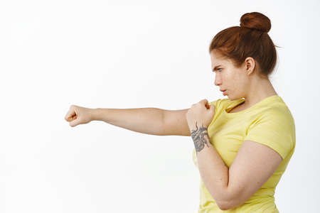 Profile portrait of serious redhead woman shadow boxing, doing box fitness workout, punching air with clenched fist, standing over white backgroundの写真素材