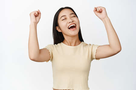 Cheerful asian girl winning, celebrating victory, making fist pump and smiling pleased, triumphing against white backgroundの写真素材