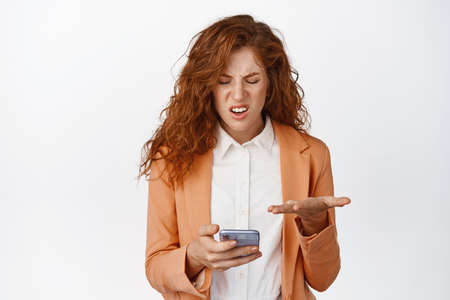 Frustrated businesswoman staring at her mobile phone and complaining, wearing suit, standing over white backgroundの写真素材