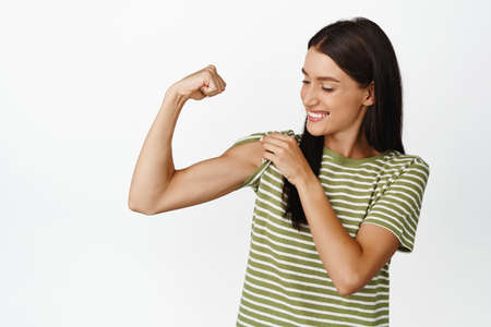 Beautiful and healthy athletic woman, showing muscles, flexing bicep and smiling, looking at her strong arm, standing over white backgroundの写真素材