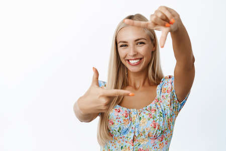Happy blond woman looking through hand frames and smiling, imaging smth, taking picture, being creative, standing over white backgroundの写真素材