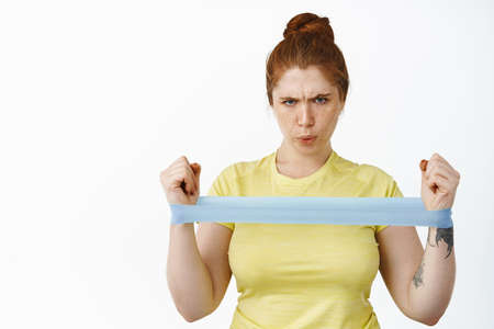 Serious redhead curvy woman doing workout with resitance band, fitness exercises on arms, stretching elastic rope, standing over white backgroundの写真素材
