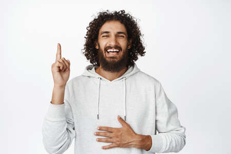 Carefree arab man laughing, pointing finger up and smiling, showing smth funny, standing in hoodie...