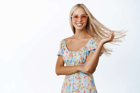 Beautiful blond woman in sunglasses, flip her long healthy hair in air, smiling and looking carefree, posing in dress over white backgroundの写真素材