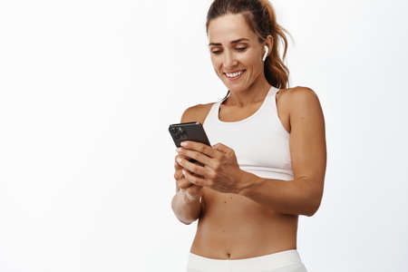 Smiling fitness woman looking at sport smartphone app, listening music wireless earphones, standing in activewear, white backgroundの写真素材